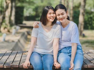 Two women sitting next to each other. One women has arm resting on shoulder of the other.